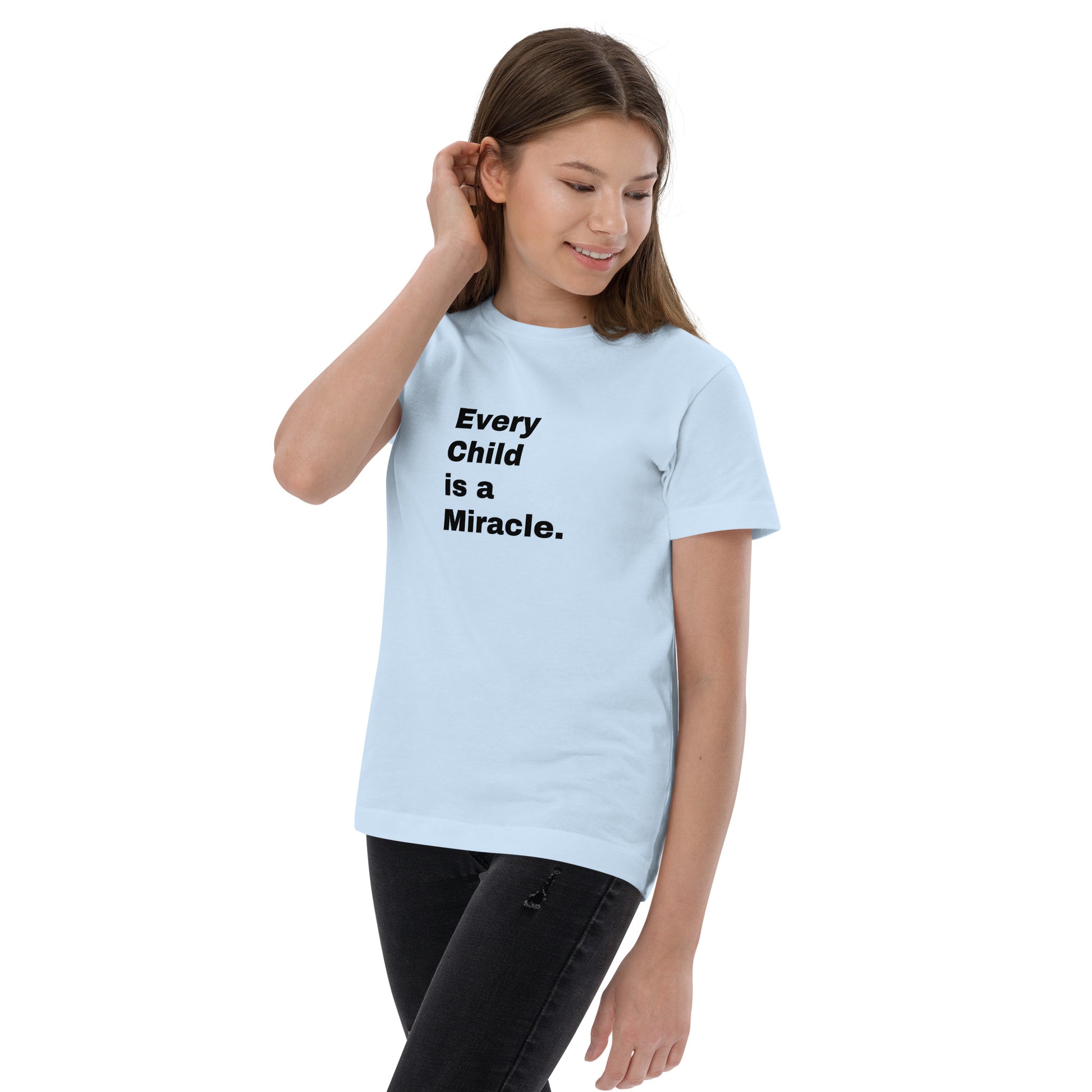 Young teen girl with long hair is tucking her hair behind her ear with one hand and has the other down by her side. She is smiling and looking down, slightly twisted, but wearing black pants and a light blue short-sleeved t-shirt that says "Every Child is a Miracle" in black text on the front.