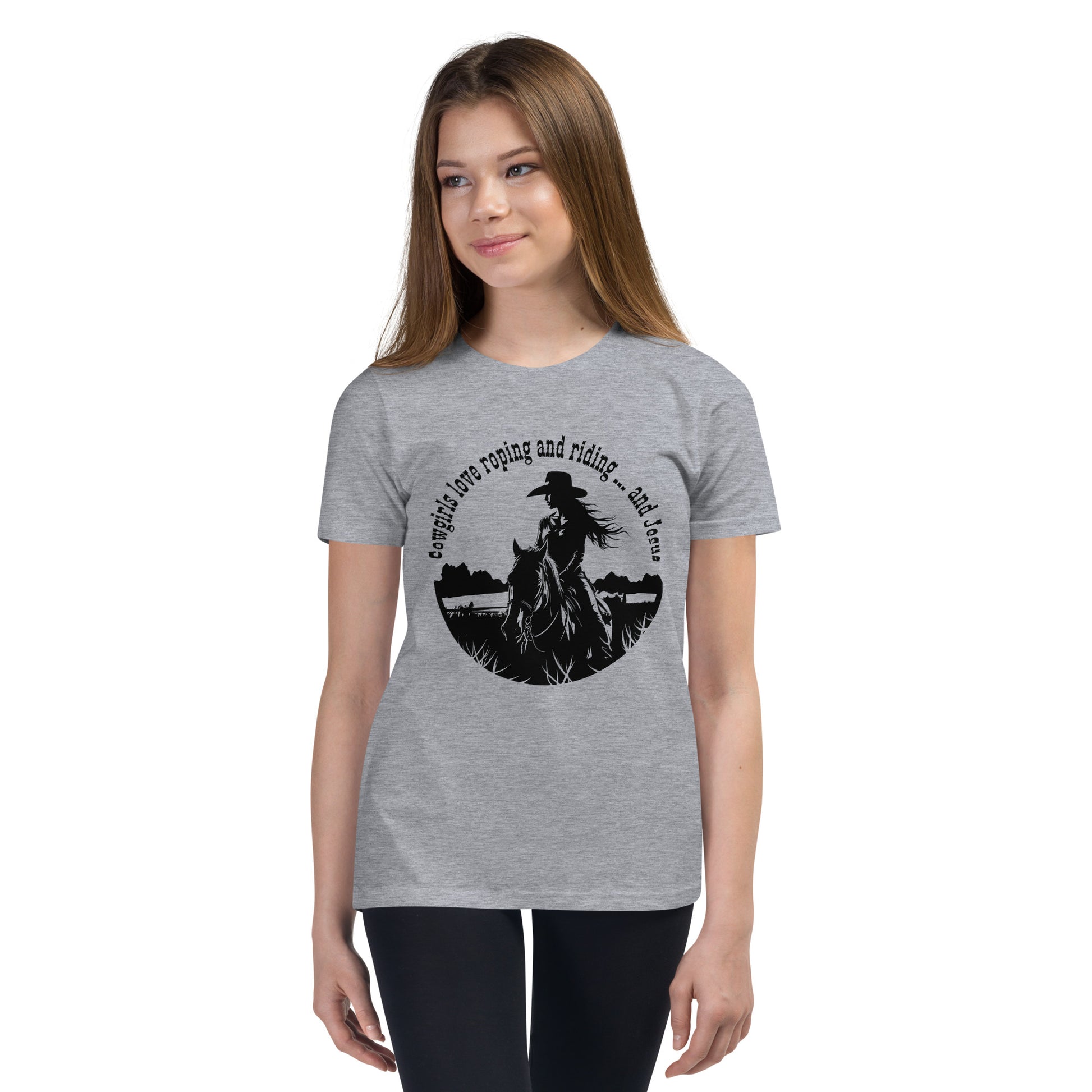 Young teen or pre-teen girl with long hair wears dark pants and a gray t-shirt that says "Cowgirls love roping and riding ... and Jesus" in black with an image of a cowgirl on horseback also in black, all cropped into a circle design.